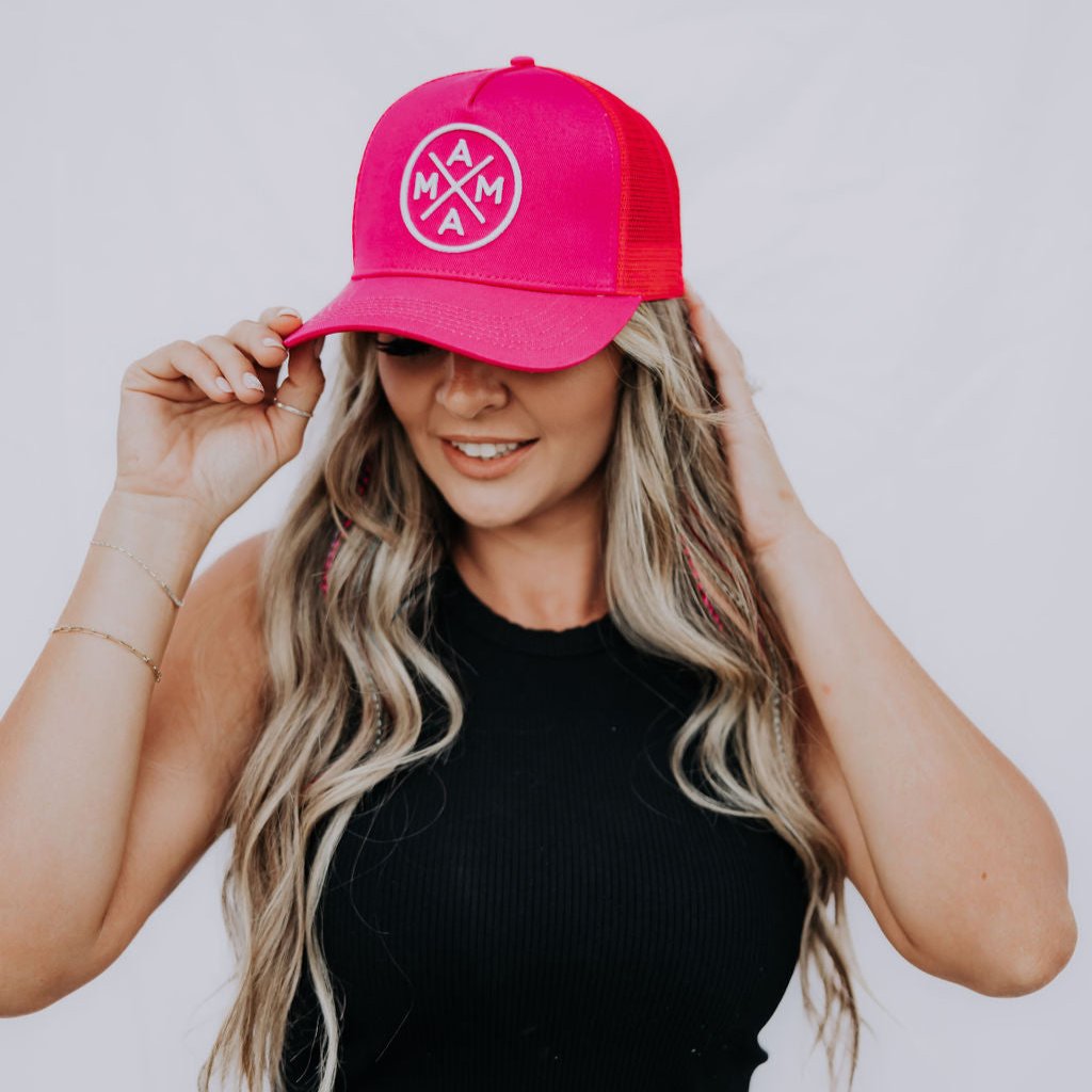 A woman with long hair is wearing a Mama X™ Premium Canvas Trucker Hat in hot pink. She pairs it with a black sleeveless top and denim jeans as she poses against a plain background, adjusting her cap with one hand and smiling.