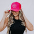 A woman with long hair is wearing a Mama X™ Premium Canvas Trucker Hat in hot pink. She pairs it with a black sleeveless top and denim jeans as she poses against a plain background, adjusting her cap with one hand and smiling.