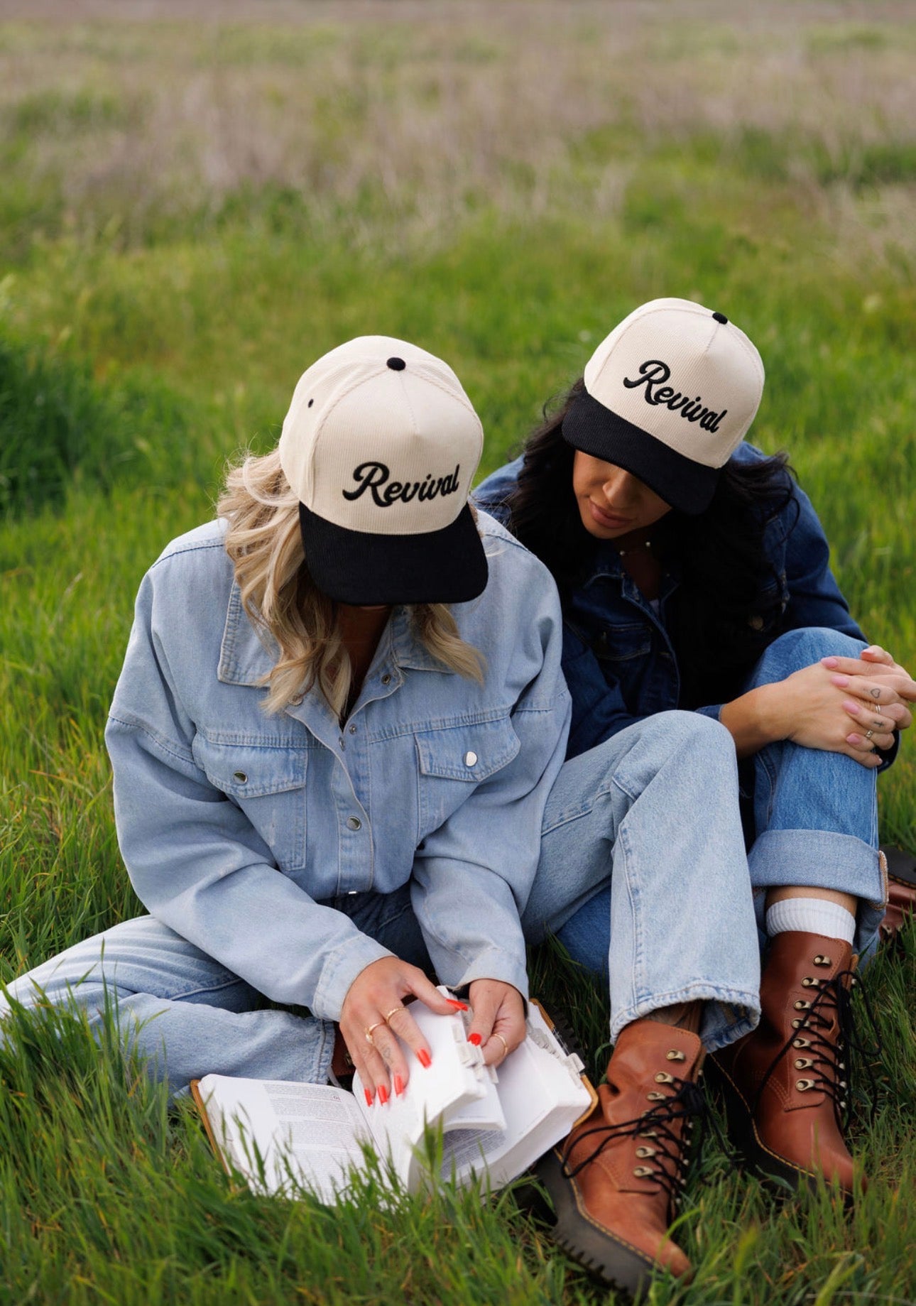 Two women sit on the grass in a field, wearing Mama X™ Brand Revival Corduroy hats and denim. One reads a faith-inspired book as the other leans in, their brown boots showing they share a moment of Rooted Revival together.