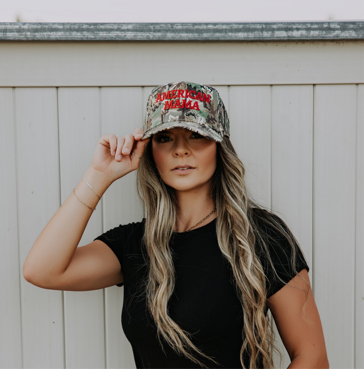 A woman with long, wavy hair wears a black t-shirt and the Mama X™ Brand American Mama Hat—a camo cap with “AMERICAN MAMA” on the front—while standing by a white wooden fence and holding the brim of her hat.