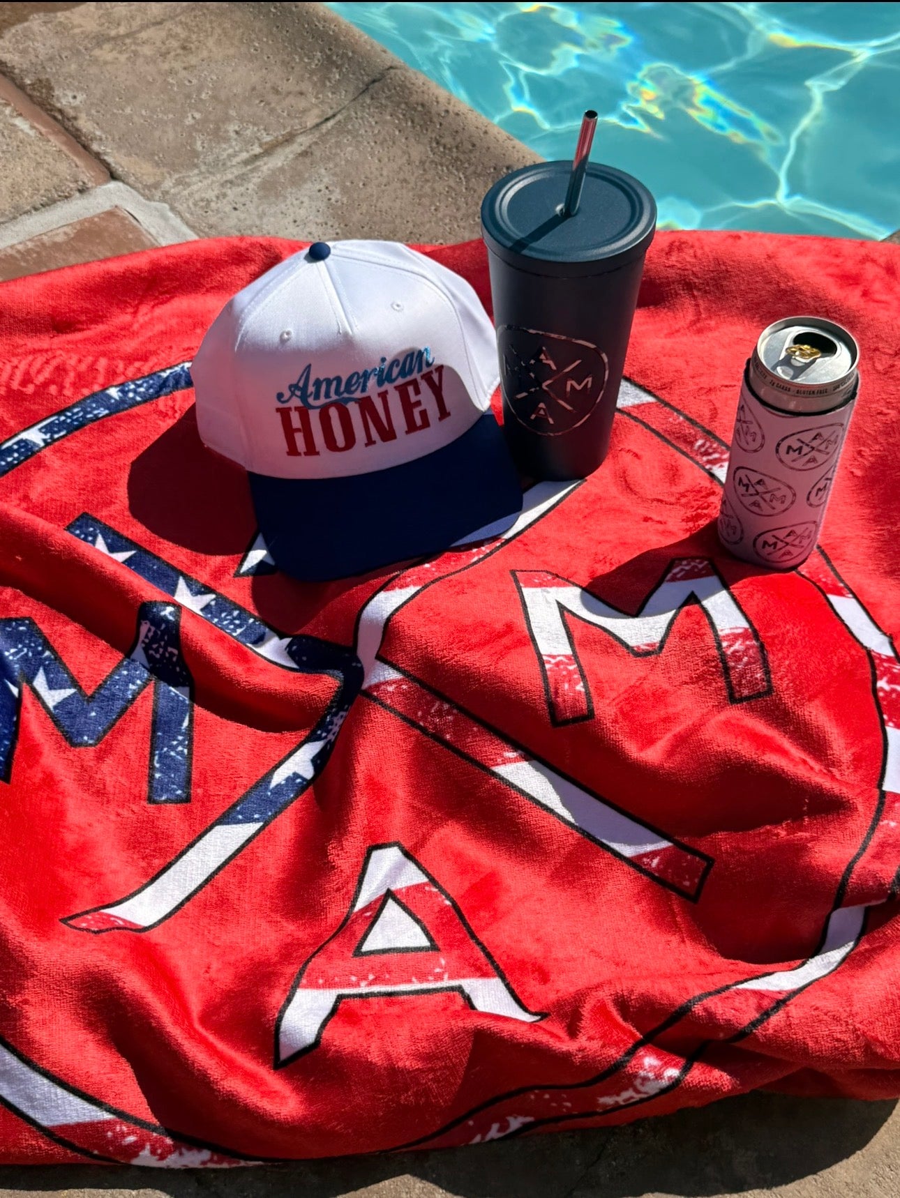 A Mama x American Flag Towel by Mama X™ Brand lies poolside, with an "American Honey" hat, a black tumbler, and a can in a pink and white koozie resting on top, all sunlit beside the sparkling pool.