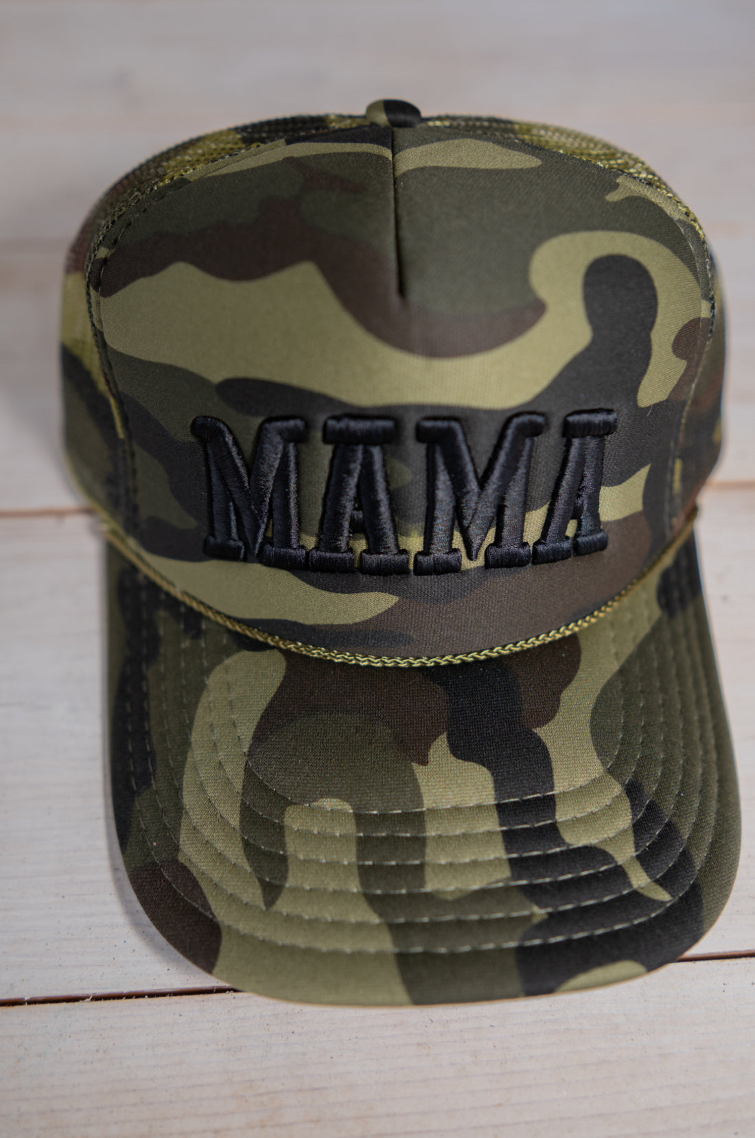 The Mama X™ Brand Black Camo Trucker baseball cap features "MAMA" in bold black embroidery on the front and is shown on a light wooden surface.