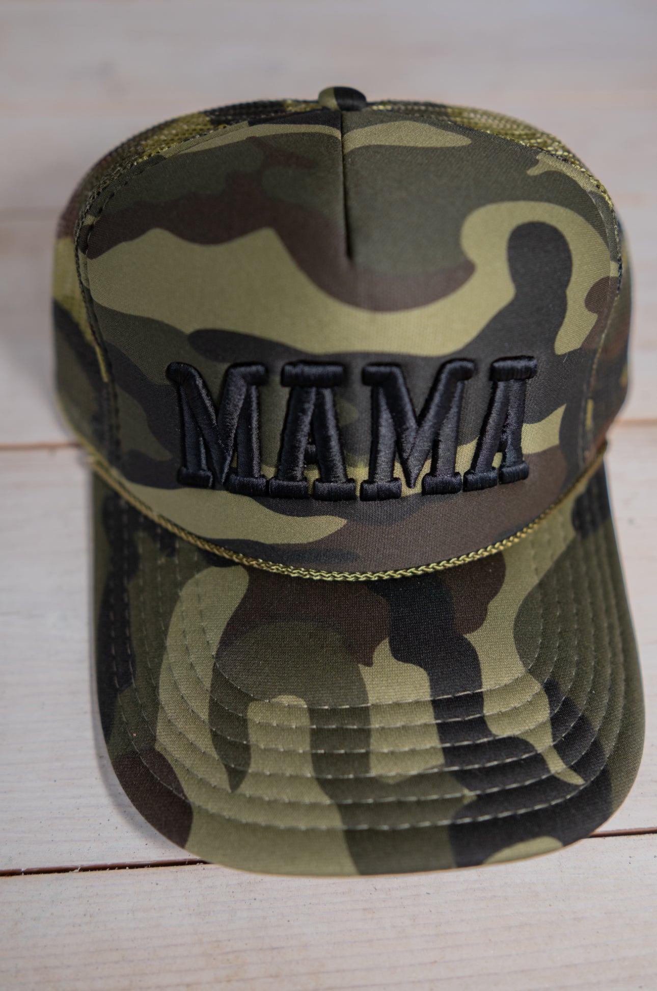 The Mama X™ Brand Black Camo Trucker baseball cap features "MAMA" in bold black embroidery on the front and is shown on a light wooden surface.