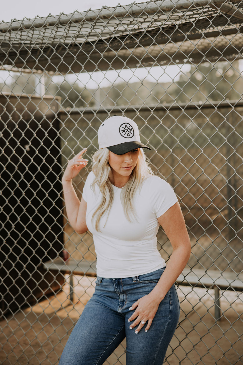A woman in a white t-shirt and blue jeans stands by a chain-link fence, looking down as she touches the brim of her Black Mama X™ Baseball Cap from Mama X™ Brand. Benches are visible in the background.