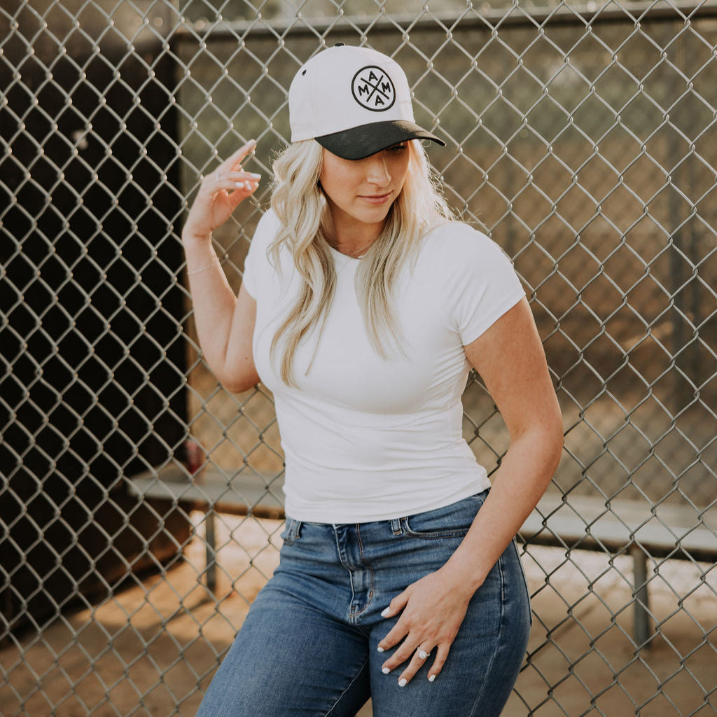 A woman in a white t-shirt and blue jeans stands by a chain-link fence, looking down as she touches the brim of her Black Mama X™ Baseball Cap from Mama X™ Brand. Benches are visible in the background.