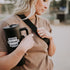 A blonde woman with a black tumbler displays a "Moms Support Moms" sticker from Mama X™ Brand. She sports a brown t-shirt and a black shoulder bag, ideal for an outing, set against a blurred outdoor scene that highlights the sticker&