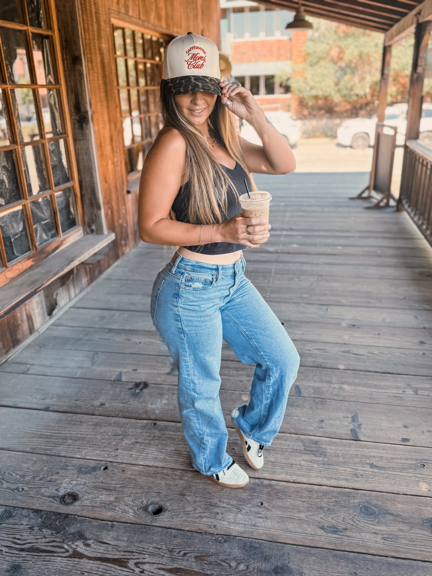 A woman wearing the Mama X™ Brand Caffeine Club Trucker cap, a black crop top, blue jeans, and sneakers smiles on a wooden porch with an iced coffee. She poses with one hand on her hat in front of a rustic building and window.