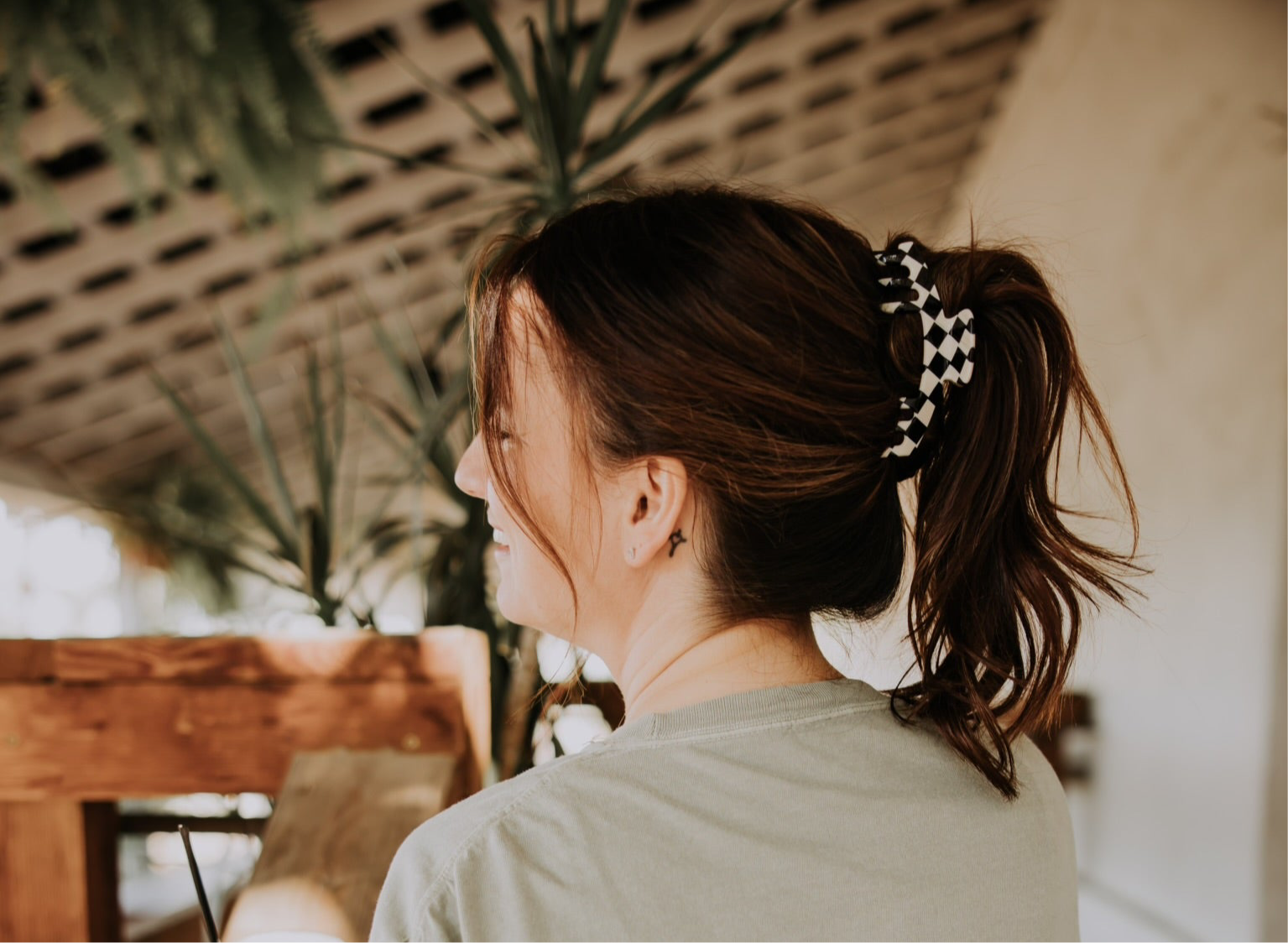 A woman with brown hair in a ponytail, secured with the Mama X™ Brand Checkered Trucker claw, smiles and looks to the side indoors, surrounded by plants and wooden beams.