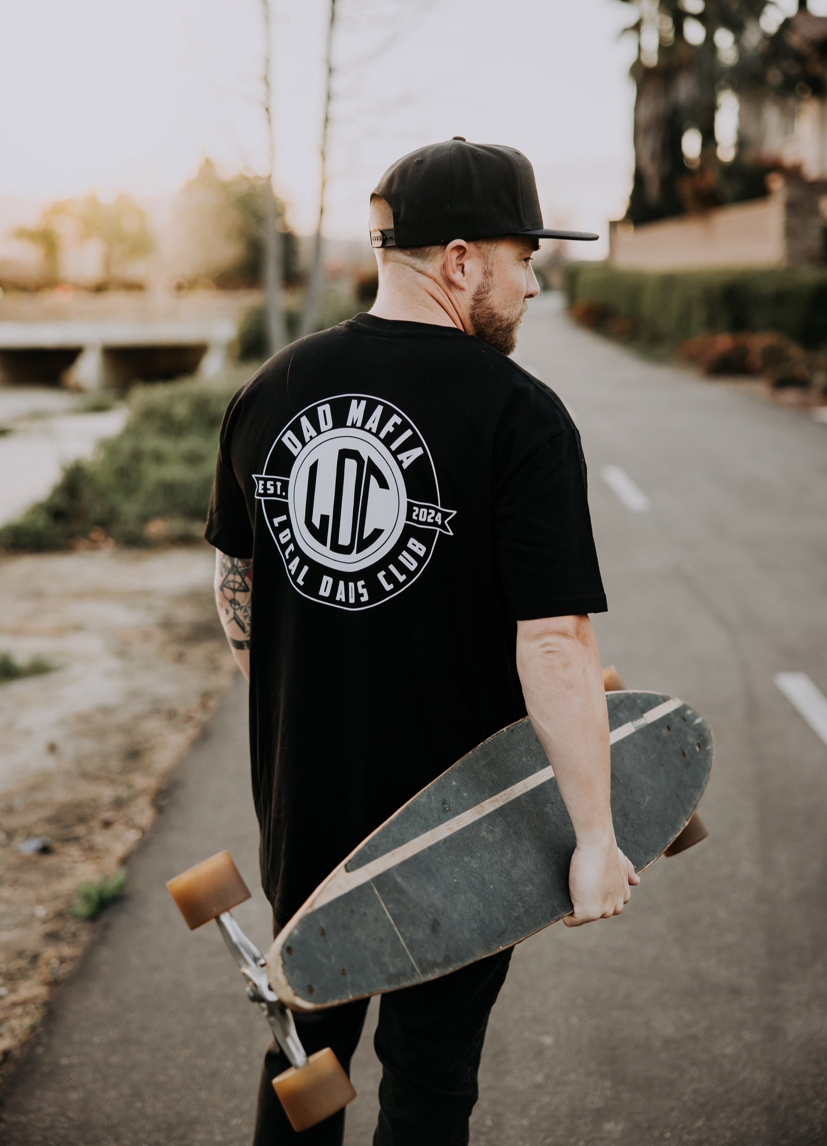 A man walking on a paved park path is holding a skateboard in his right hand. He is wearing a black baseball cap and the Dad Mafia T-Shirt from the Mama X™ Brand, made of 100% ringspun cotton, with 