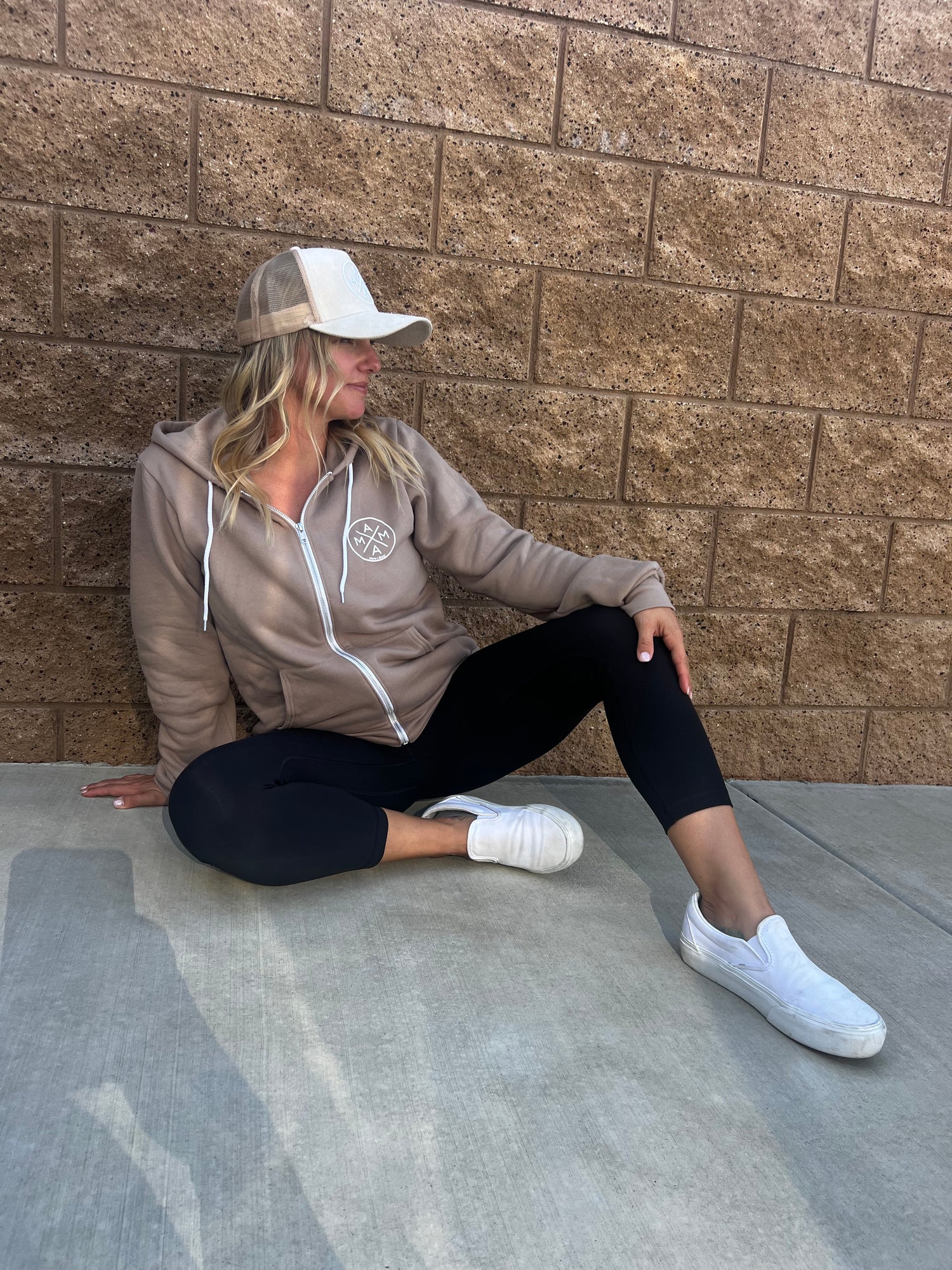 A person wearing a brown Mama X™ Zip Up Sweatshirt from the Mama X™ Brand, black leggings, and white sneakers is sitting on the ground against a textured brick wall. They are looking to their right with one leg bent and the other stretched out.