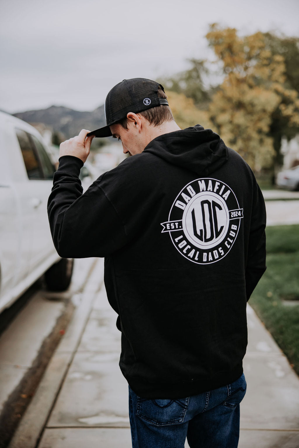 A man wearing a black Dad Mafia Hoodie from Mama X™ Brand and a black cap stands on a sidewalk, facing away from the camera. The heavy-weight hoodie features a fleece-lined hood with a circular logo that reads 