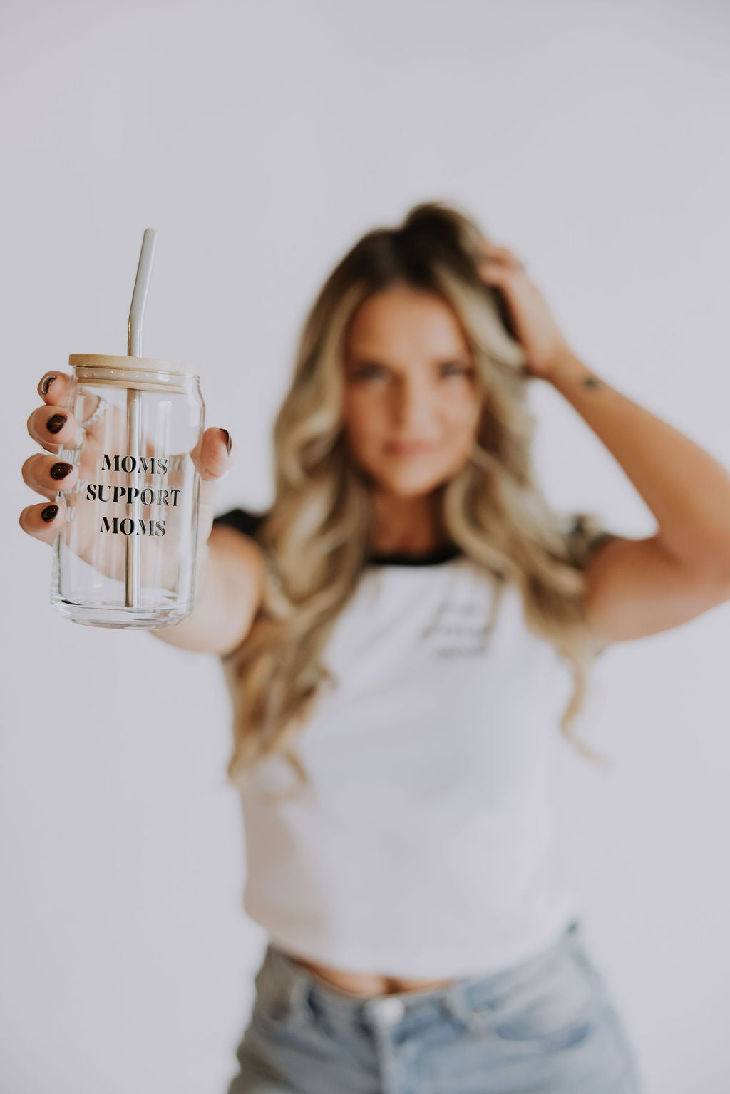 A woman with long blonde hair holds out the 16 oz Mama X™ Brand Moms Support Moms Glass mug with a straw. She wears a white t-shirt and jeans, standing against a softly blurred background.