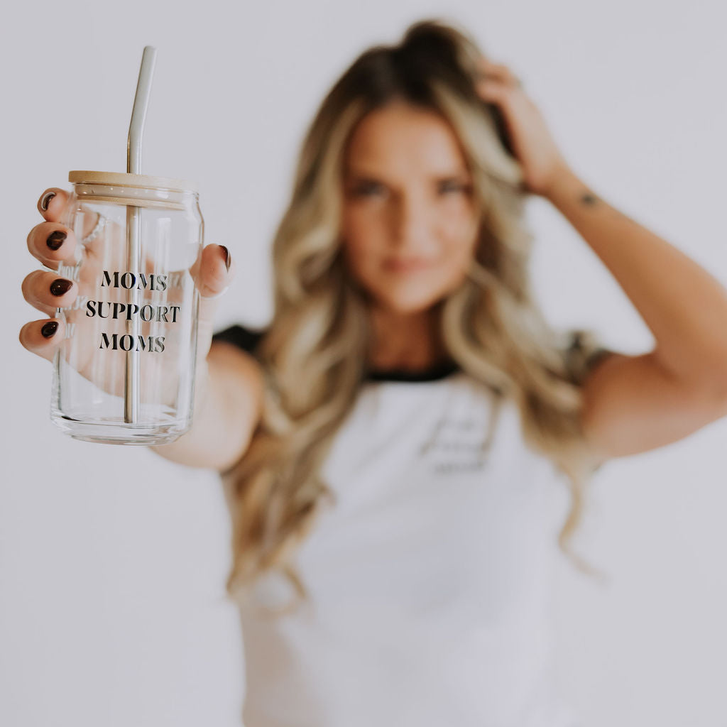 A woman with long blonde hair holds out the 16 oz Mama X™ Brand Moms Support Moms Glass mug with a straw. She wears a white t-shirt and jeans, standing against a softly blurred background.