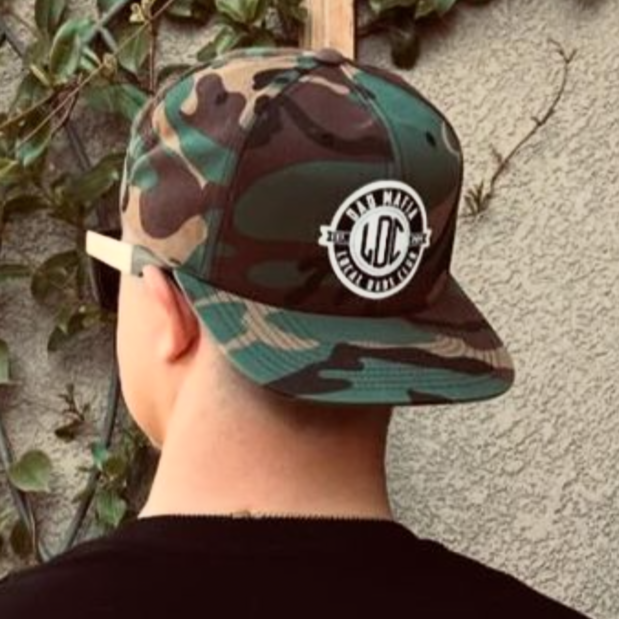 A person with their back to the camera is wearing a Dad Mafia Camo Snapback from the Mama X™ Brand. The cap features an embroidered logo on the back with the letters 