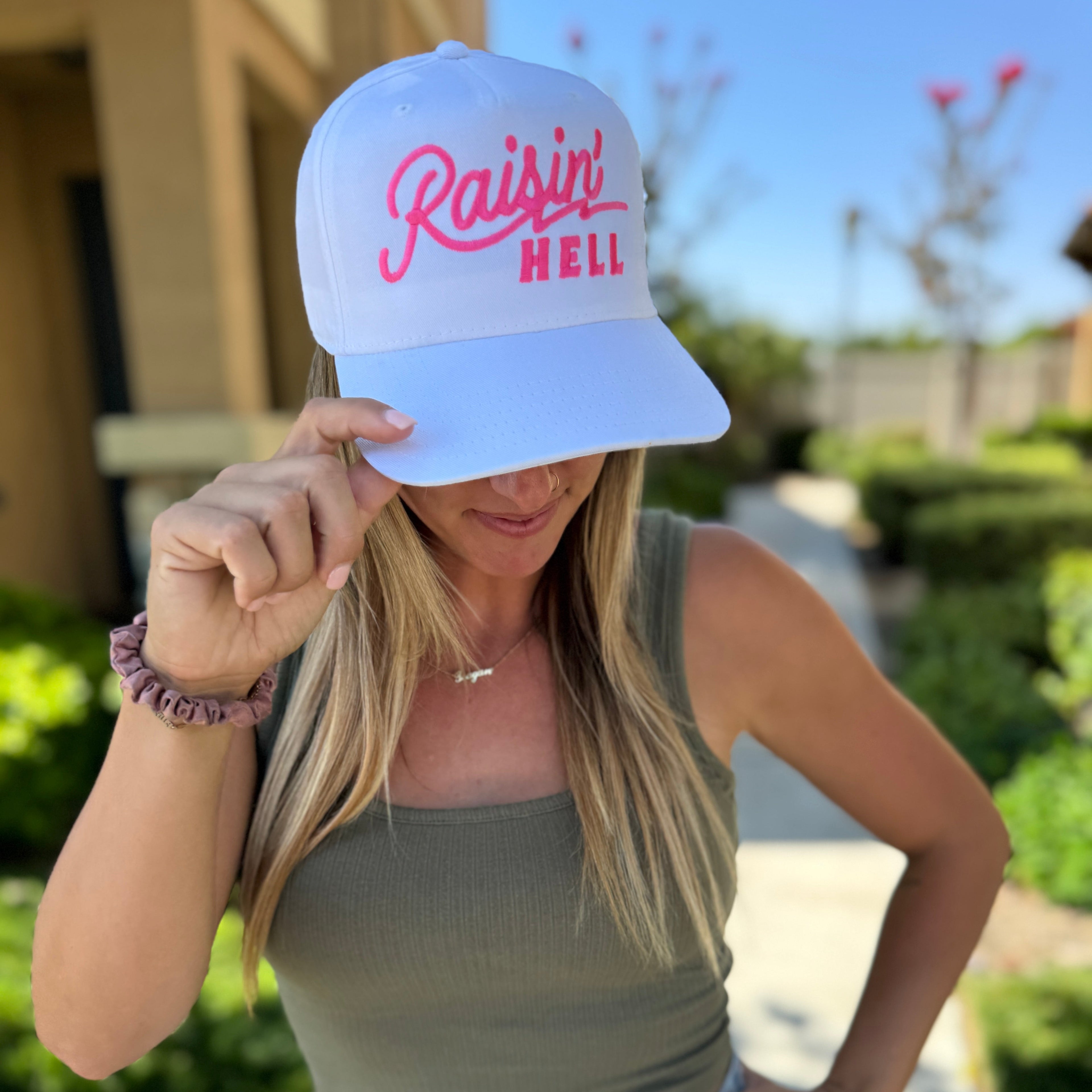 A woman in a green tank top and denim shorts stands outside, playfully holding her Mama X™ Brand Raisin’ Hell White Trucker cap—perfectly capturing that raisin&