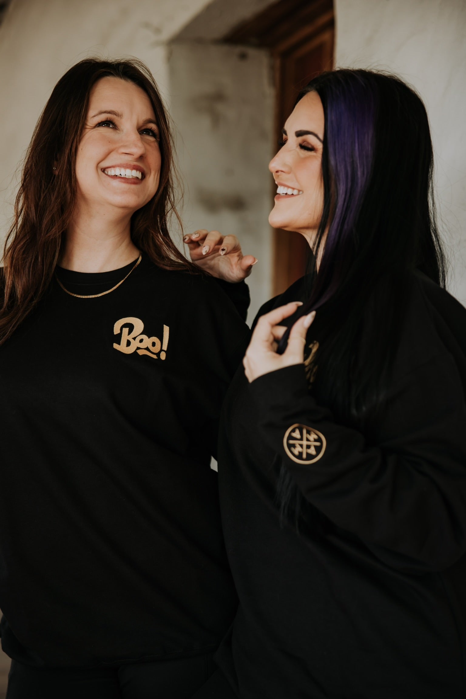 Two women in a casual setting wear Mama X™ Brand tops—one sports the Boo Crewneck with "Boo!" in gold lettering, while the other has a black shirt featuring a circular gold symbol on the sleeve. Both appear happy and relaxed.