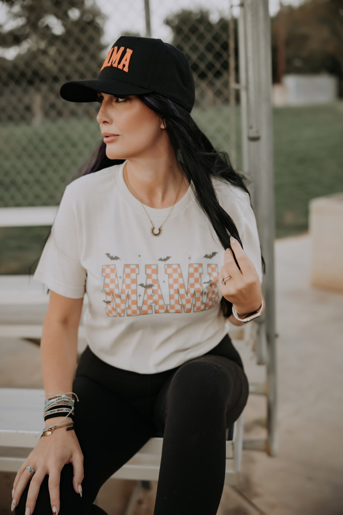 Woman with long dark hair wears a black cap and the Mama X™ Brand Mama Halloween Tshirt with black leggings and bracelets, sitting on an outdoor bench by a chain-link fence, looking to the side.