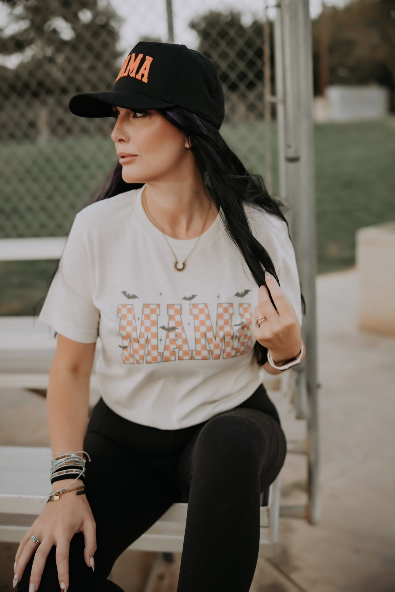 Woman with long dark hair wears a black cap and the Mama X™ Brand Mama Halloween Tshirt with black leggings and bracelets, sitting on an outdoor bench by a chain-link fence, looking to the side.