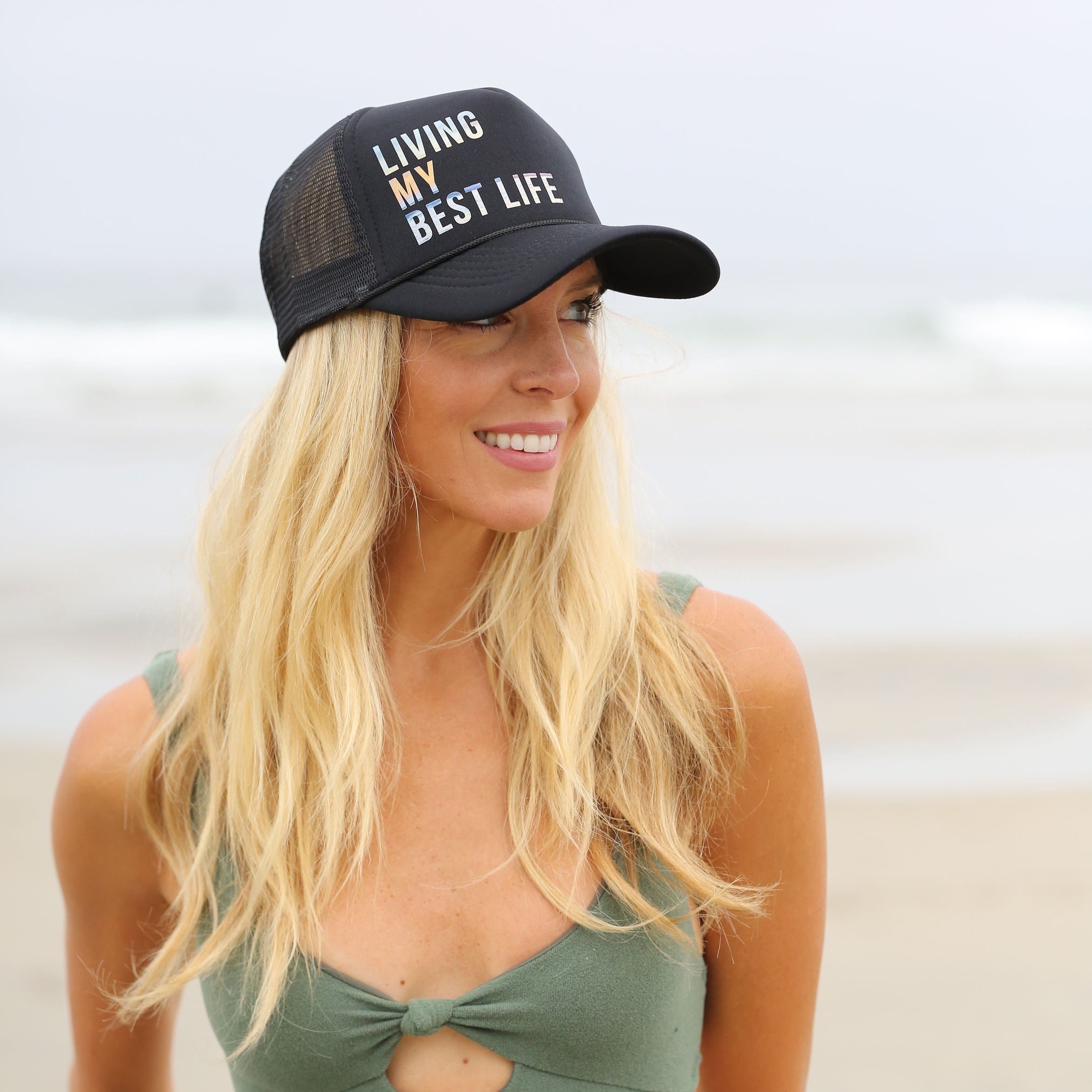 A blonde woman stands on a sandy beach, smiling and gazing to the side. She is wearing a green sleeveless top and the 