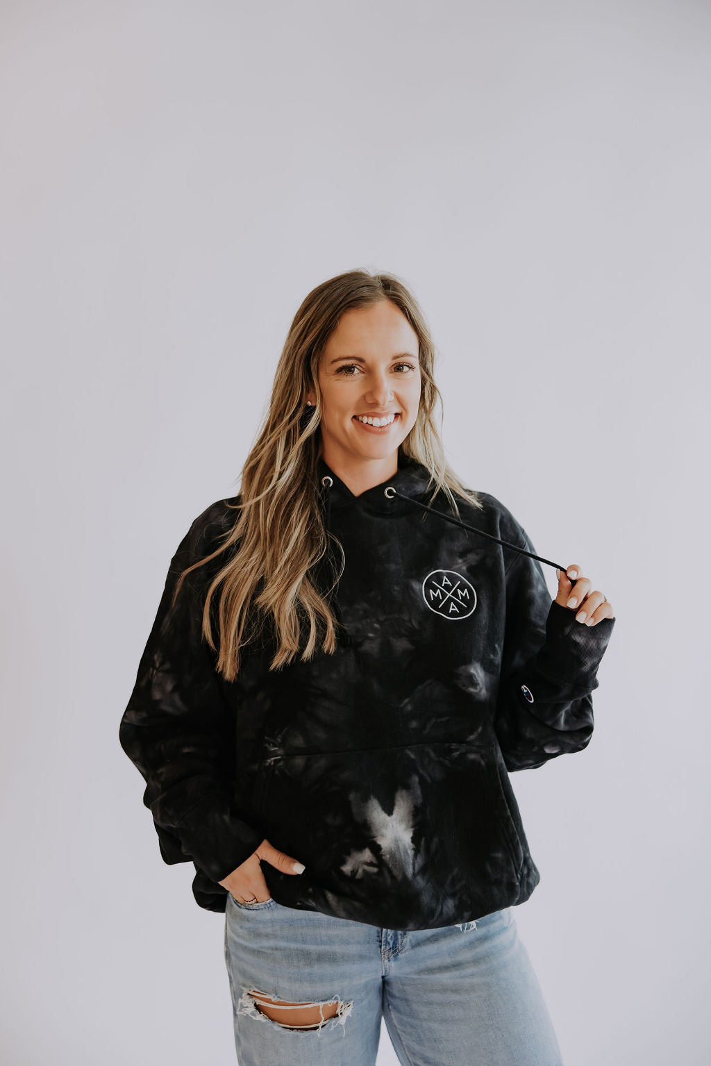 A smiling woman with long, wavy blonde hair wears a trendy Mama X™ Brand Mama X Embroidered Hoodie and ripped light blue jeans, standing against a plain light background. She holds one hoodie drawstring in her right hand.