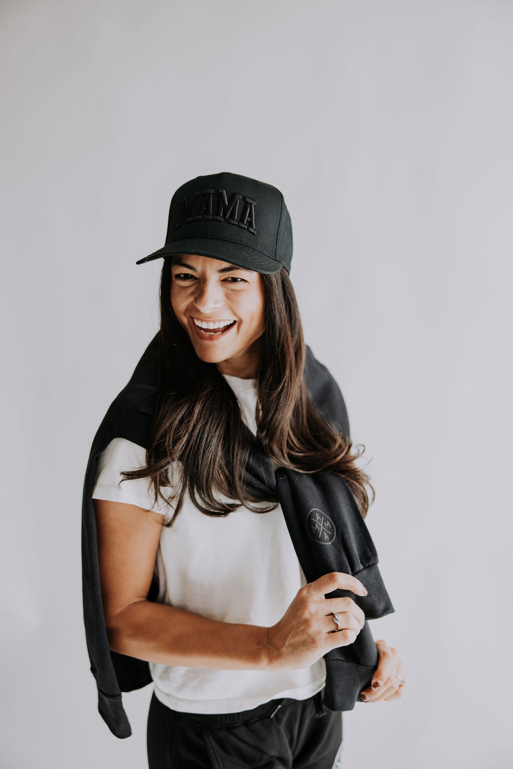 A smiling woman with long brown hair wears the Mama X™ Brand Mama black on black Trucker Hat, a black baseball cap with 3D embroidery, paired with a white t-shirt and a black sweatshirt over her shoulders against a plain light background.