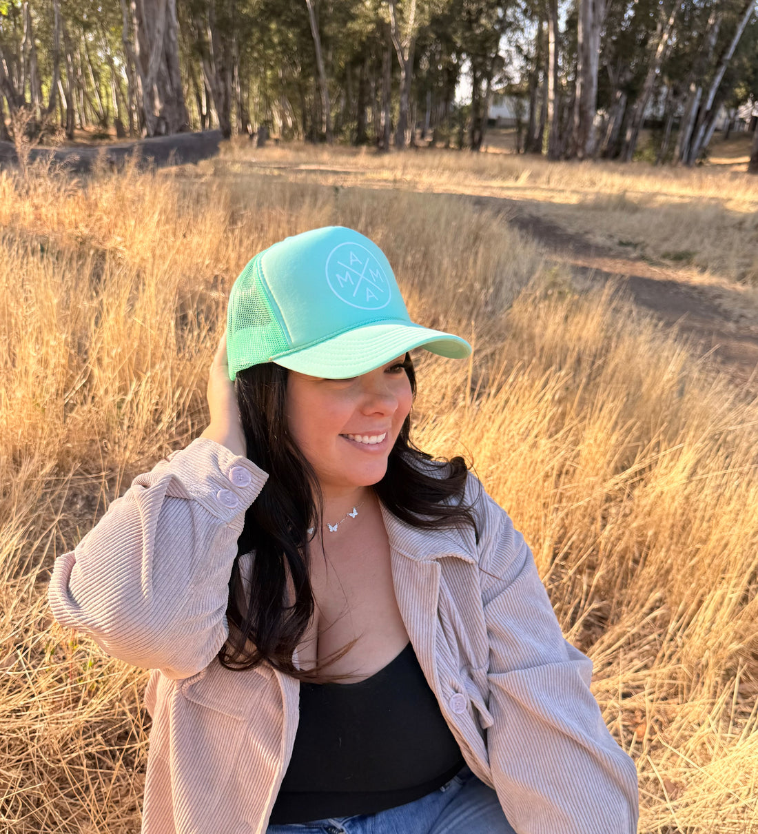 A woman smiles in a dry grassy field under bright sunlight, wearing the Mama X™ Brand Teal foam Mama X Trucker hat, a beige jacket, and a black top. Tall trees stand in the background.