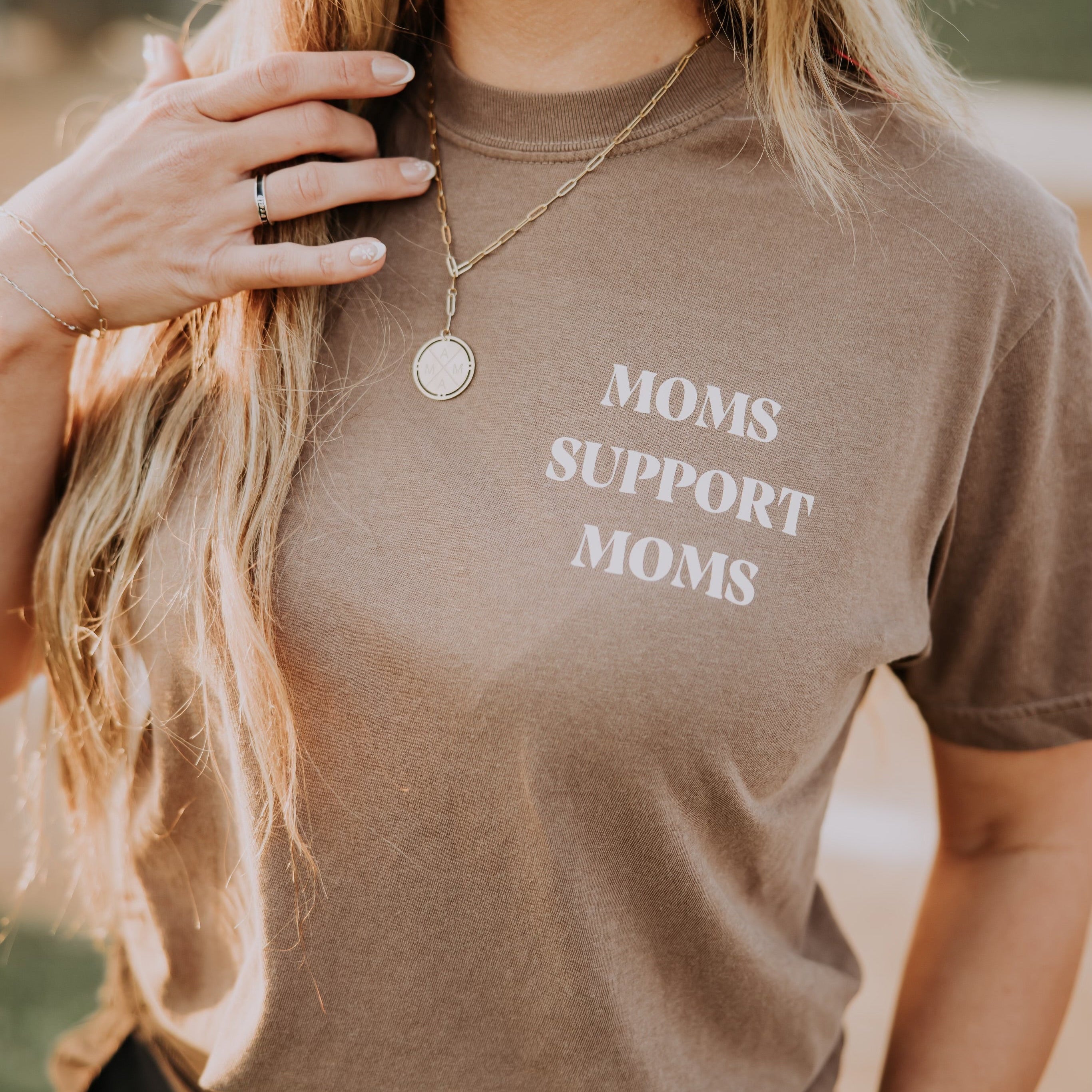 A person with long, blonde hair wears a garment-dyed brown "Moms Support Moms" T-shirt from Mama X™ Brand, featuring a necklace and a ring on their left hand. The blurred background suggests an outdoor setting, capturing the essence of a supportive mama.