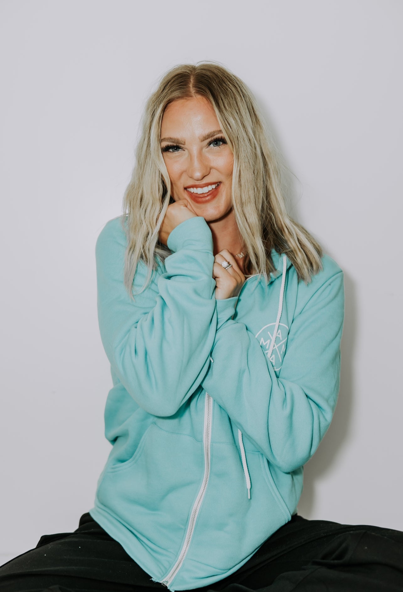 A smiling woman with blonde hair sits against a plain background. She is wearing the Mama X™ Zip Up Sweatshirt in teal from Mama X™ Brand and black pants. Her hands are gently touching her face, and she appears to be in a relaxed and cheerful mood.