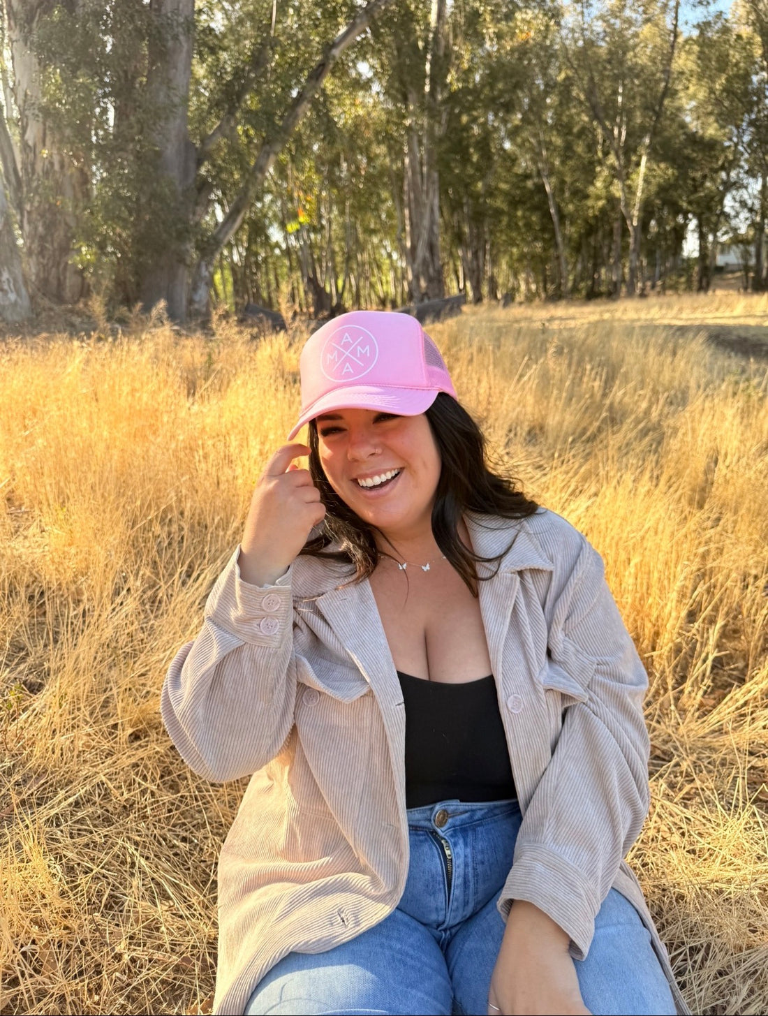 A woman in a light jacket, black top, and blue jeans smiles in tall golden grass, holding the brim of her Mama X™ Brand Pink foam Mama X Trucker hat, with sunlit trees in the background—a perfect snapshot of mom life.