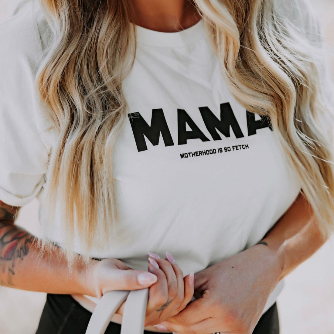 A woman with long, wavy blonde hair wears the Mama X™ Brand "Motherhood Is So Fetch" crop top, a relaxed boxy white t-shirt in 100% combed cotton featuring “MAMA” and “Motherhood is so fetch,” while holding light-colored straps.