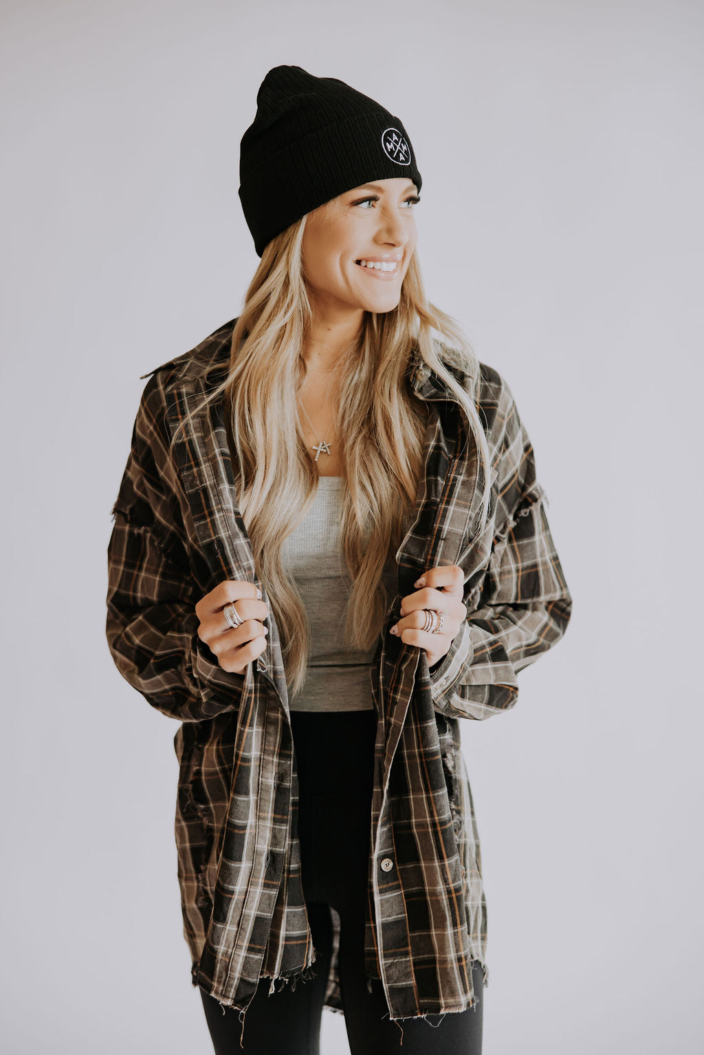 A woman with long blonde hair smiles and looks to the side against a plain, light background, wearing the Mama Cuffed Beanie by Mama X™ Brand with a plaid flannel over a gray top and black pants.