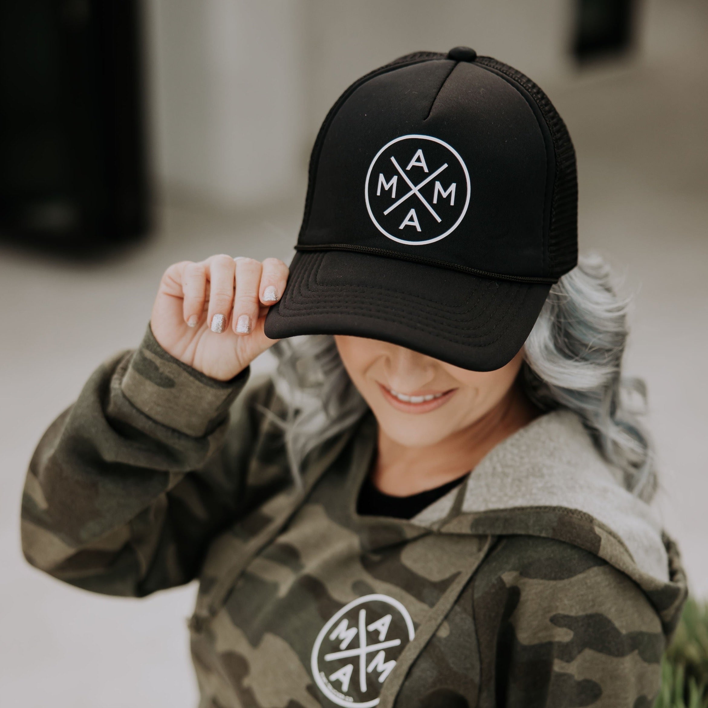 A person with long hair, wearing a black Mama X™ Foam Trucker Hat and a camouflage hoodie, both featuring a circular logo with the letters "MAMA" arranged in a cross pattern, is tilting the hat slightly forward while looking down.