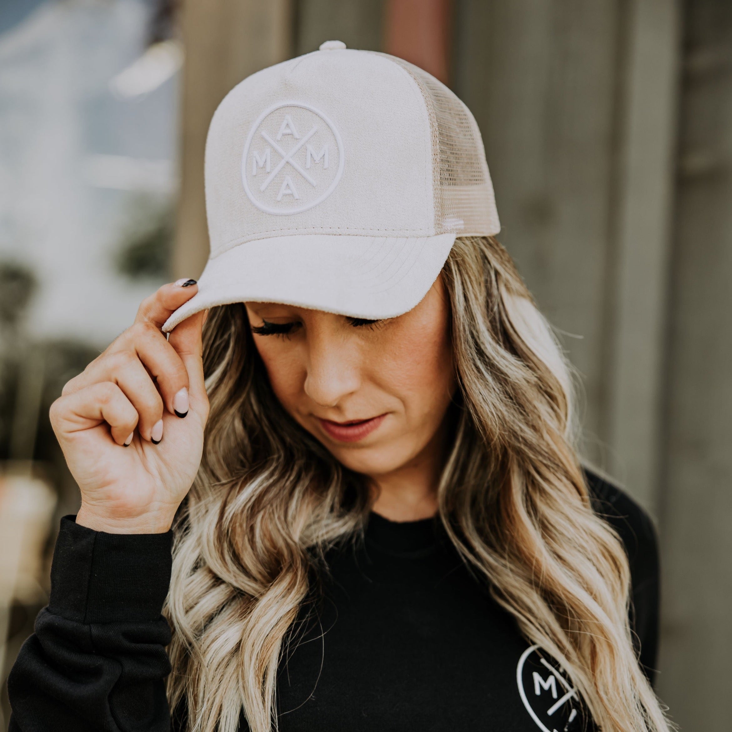 A person with long wavy hair wears a beige Mama X™ Light Suede Trucker Hat featuring the letters 