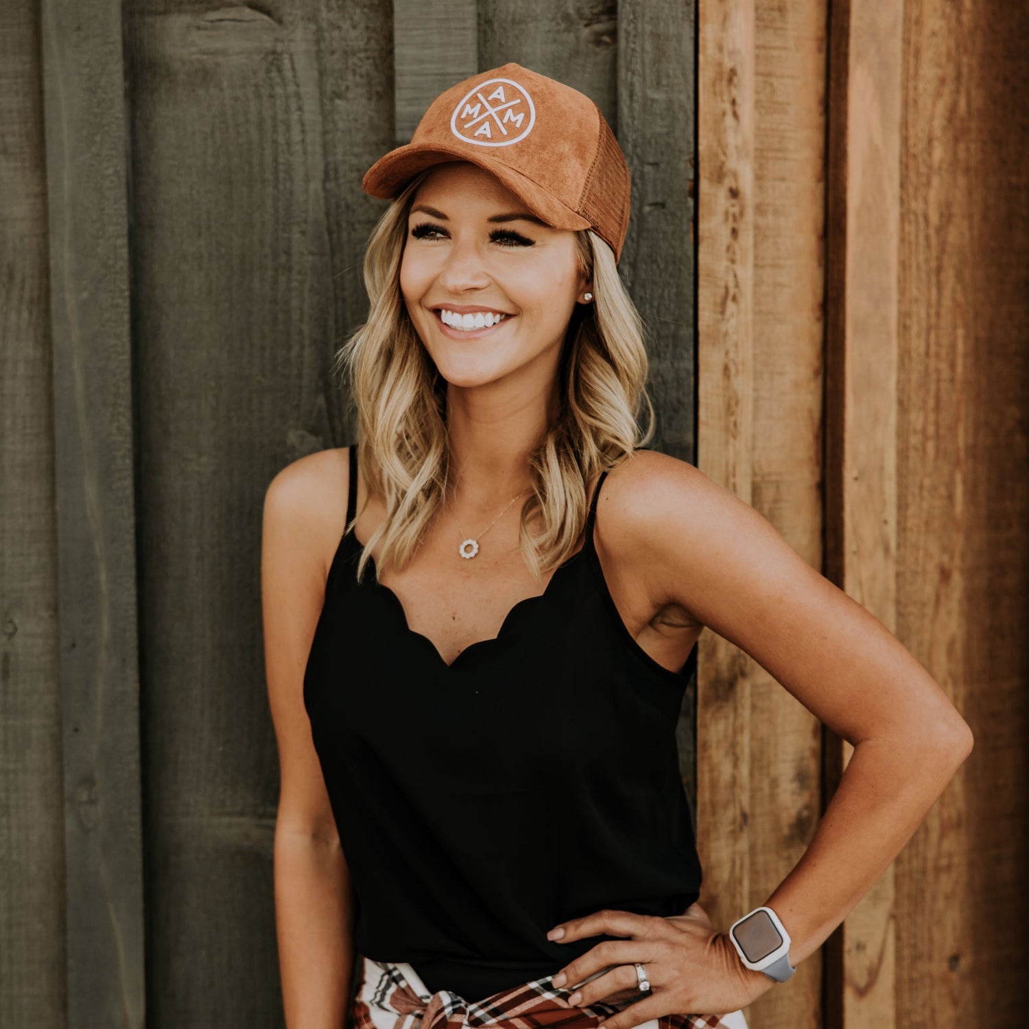 A smiling person stands against a wooden wall, wearing a black scalloped-edge tank top, a Mama X™ Trucker Hat - Brown Suede by Mama X™ Brand, and a watch on their left wrist. Their blonde hair is styled in loose waves, completing the perfect fall outfit with a plaid shirt tied around their waist.