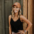 A smiling person stands against a wooden wall, wearing a black scalloped-edge tank top, a Mama X™ Trucker Hat - Brown Suede by Mama X™ Brand, and a watch on their left wrist. Their blonde hair is styled in loose waves, completing the perfect fall outfit with a plaid shirt tied around their waist.