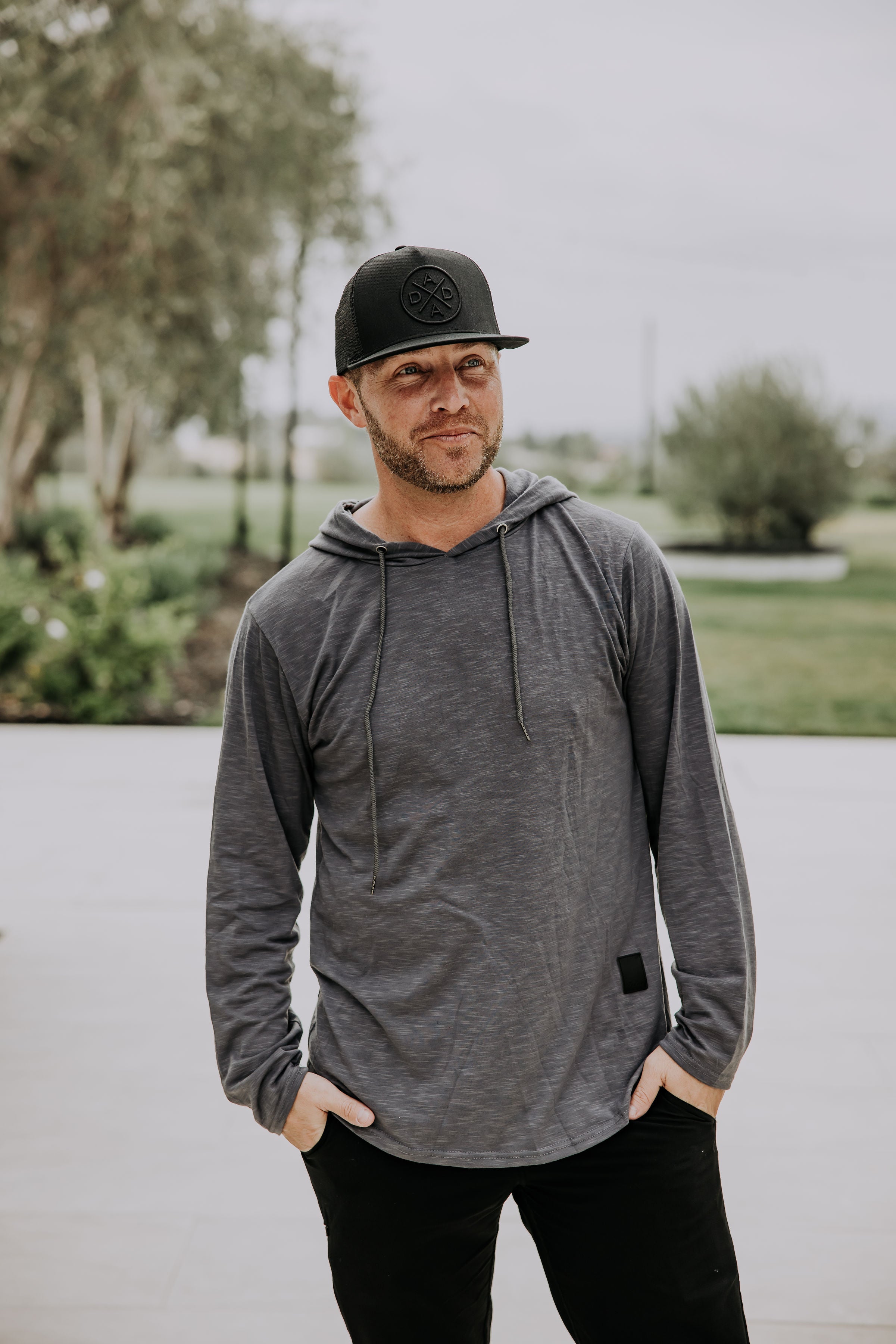 A man wearing a gray hoodie and a Dada X™ Blackout Premium Trucker Hat with a stitched 3D logo from Mama X™ Brand stands outdoors with his hands in his pockets. He has a slight beard and is looking to the side with a contemplative expression. The background features greenery and trees, providing a natural, serene setting.