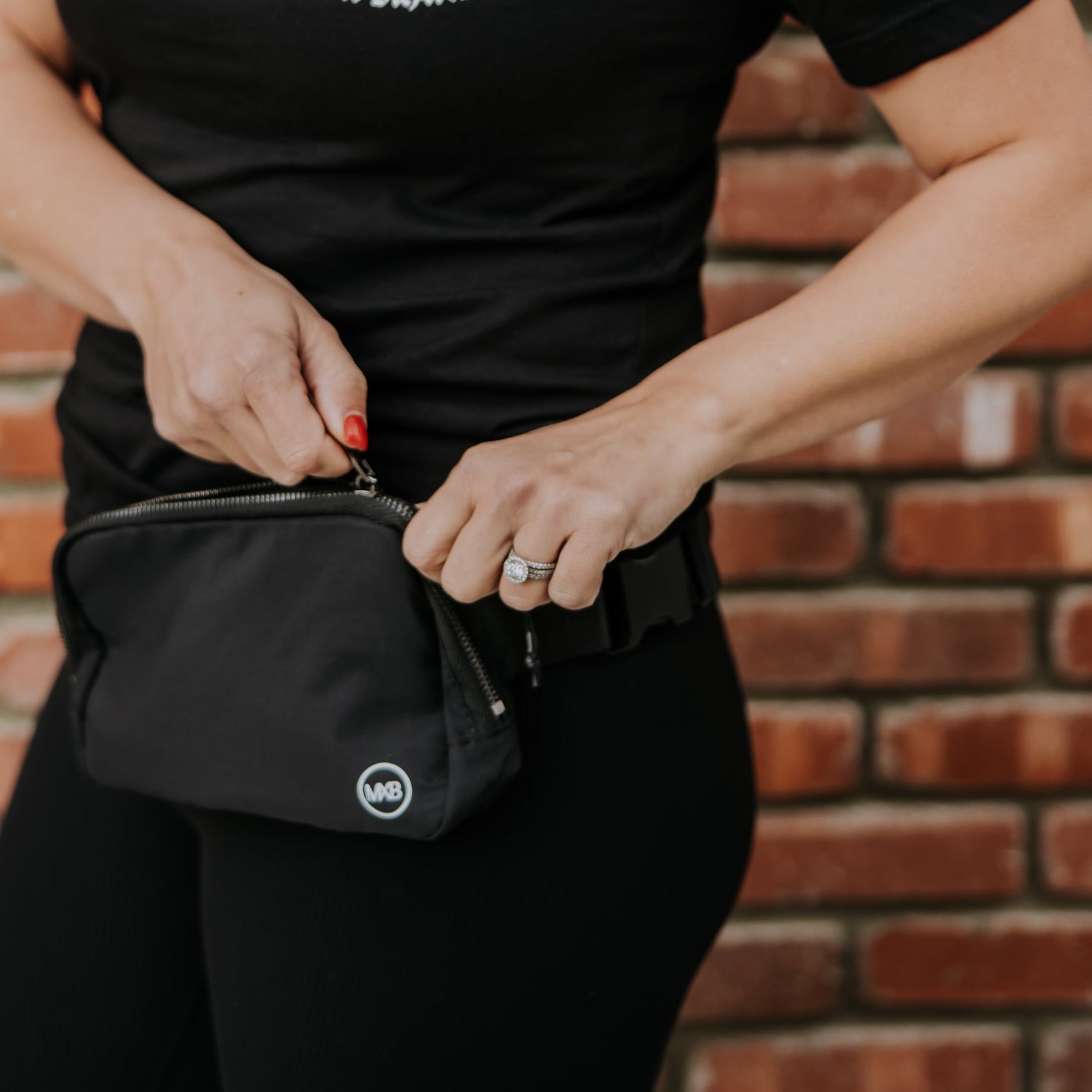 A person wearing a black shirt and leggings opens a Mama X™ Brand MXB Crossbody Bag with an MB logo on the lower right corner in front of a brick wall. The person&