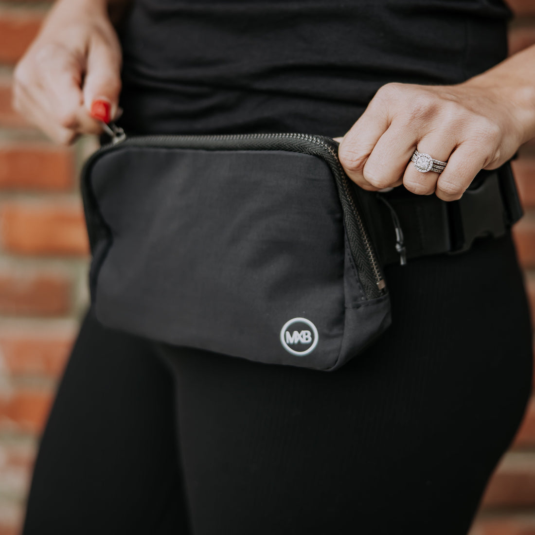 A person wearing black pants and a black shirt is adjusting the MXB Crossbody Bag from Mama X™ Brand—an absolute closet must-have—around their waist. They have a ring on their finger. The bag, which can also be worn as a fanny pack, features the logo with the letters "MB." A brick wall is partially visible in the background.