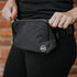 A person wearing black pants and a black shirt is adjusting the MXB Crossbody Bag from Mama X™ Brand—an absolute closet must-have—around their waist. They have a ring on their finger. The bag, which can also be worn as a fanny pack, features the logo with the letters "MB." A brick wall is partially visible in the background.