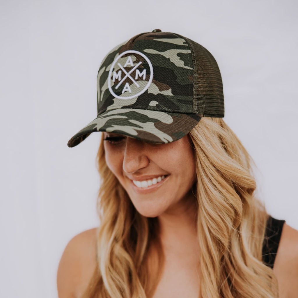 A person with long blonde hair is smiling while wearing the Mama X™ Brand's Premium Canvas Trucker Hat in camo, which features the iconic crossed arrows logo. Paired with a black tank top, this stylish look pops against the plain white background.