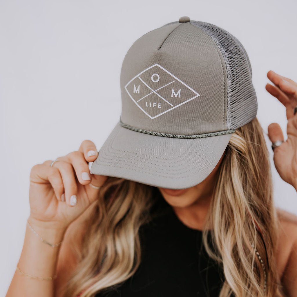 A woman with long, wavy blonde hair wearing a high-quality "Mom Life" foam trucker hat from Mama X™ Brand adjusts its brim. She's dressed in a black sleeveless top against a plain background, showcasing the adjustable snap on her stylish gray Mom Life Foam Hat - Gray.