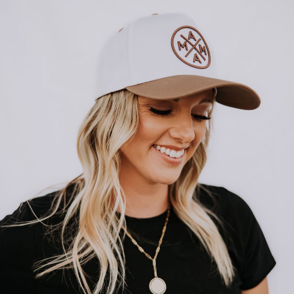 A person with long, wavy blonde hair is smiling while wearing a black t-shirt and a Brown Mama X™ Baseball Cap. The cap showcases the Mama X™ logo with the letters "MAMA." They are also adorned with a gold coin necklace against a plain white backdrop.