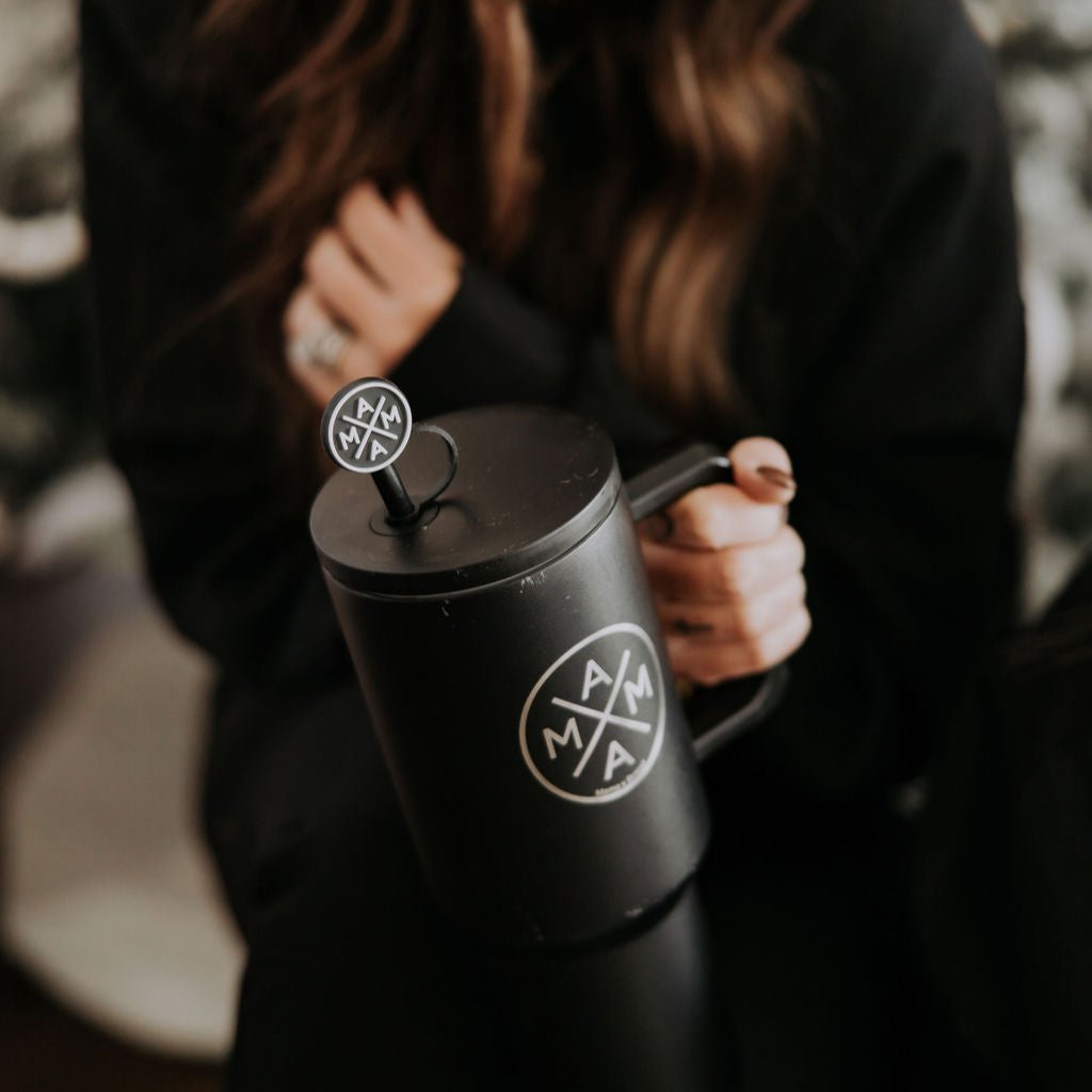 A person is holding a black coffee mug adorned with the Mama X™ Brand logo in white, stylized letters inside a circle. They are wearing a dark outfit and have opted for minimalistic accessories. Their face is not visible, and the background remains slightly blurred.