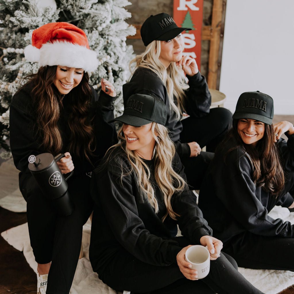 Four women in matching black outfits and "MAMA" hats with vibrant accents gather by a snow-dusted Christmas tree. One dons a Santa hat, and another's mug features the Mama X™ Straw Topper by the Mama X™ Brand. They share laughter and holiday spirit, with a Christmas sign displayed in the background.