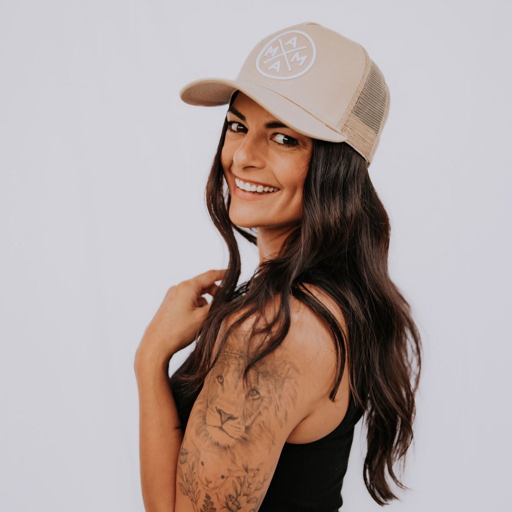 A woman with long dark hair and a tattooed arm smiles while posing in a black tank top and denim jeans. She wears the Mama X™ Premium Canvas Trucker Hat in tan, its subtle logo adorning her cap as she faces sideways against a plain white background.