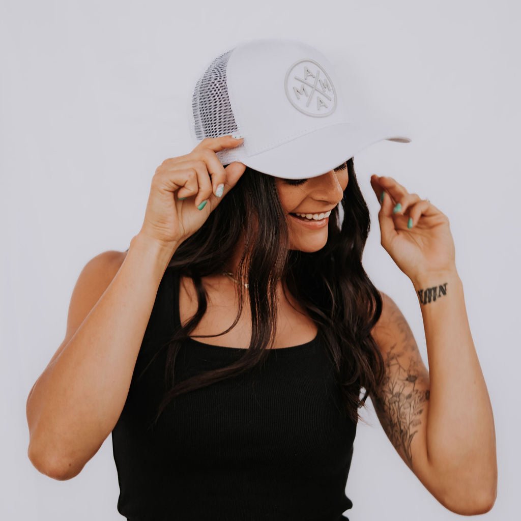 A woman with long dark hair and tattoos on her right arm smiles while adjusting her Mama X™ Premium Canvas Trucker Hat in white and gray. She is wearing a black tank top and jeans, set against a plain white background.