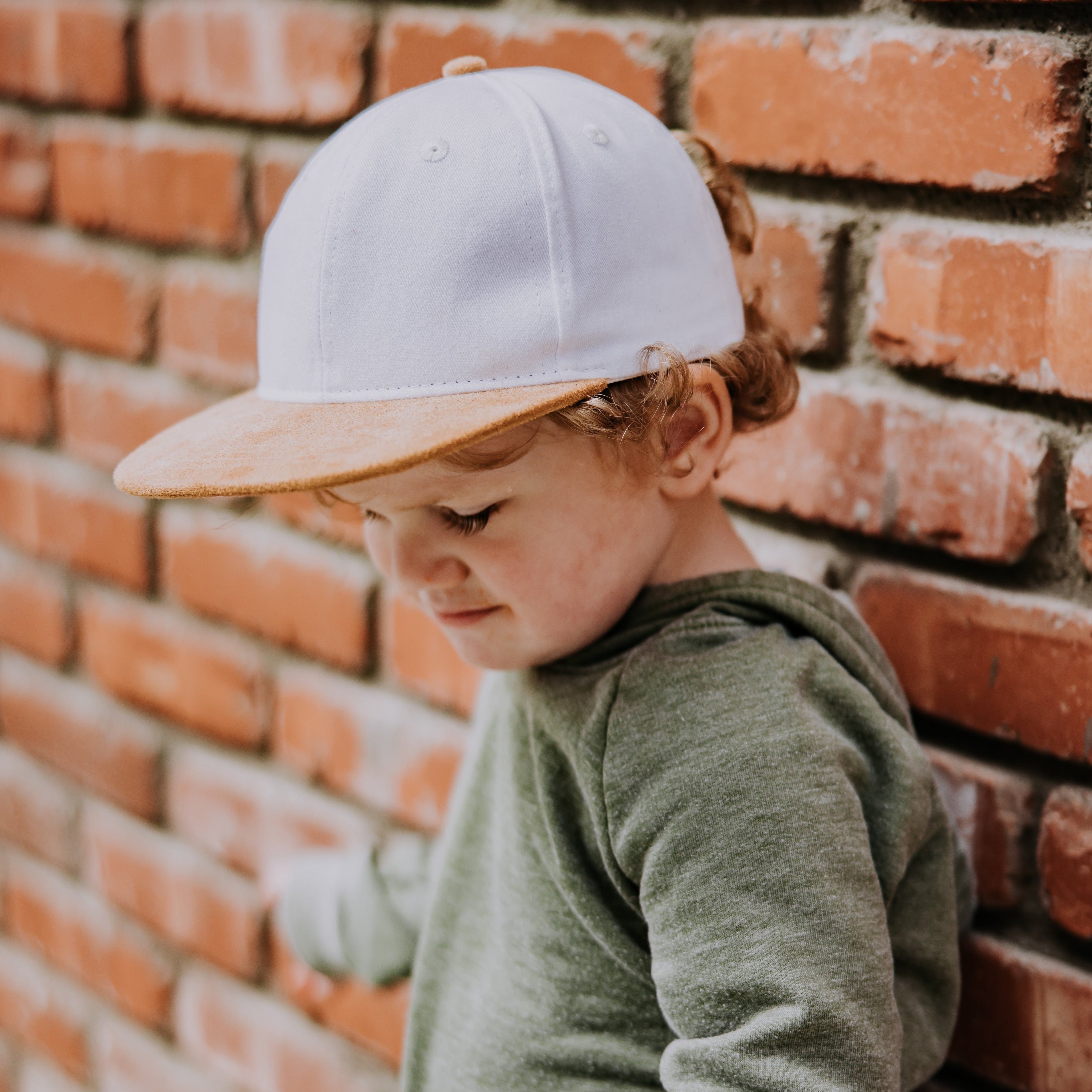 A young child with curly hair is wearing a White with Brown Suede Bill Trucker hat from the Mama X™ Brand and a green sweater. The child, dressed in toddler & youth sizes, is standing against a brick wall, looking down with a slight smile on their face.