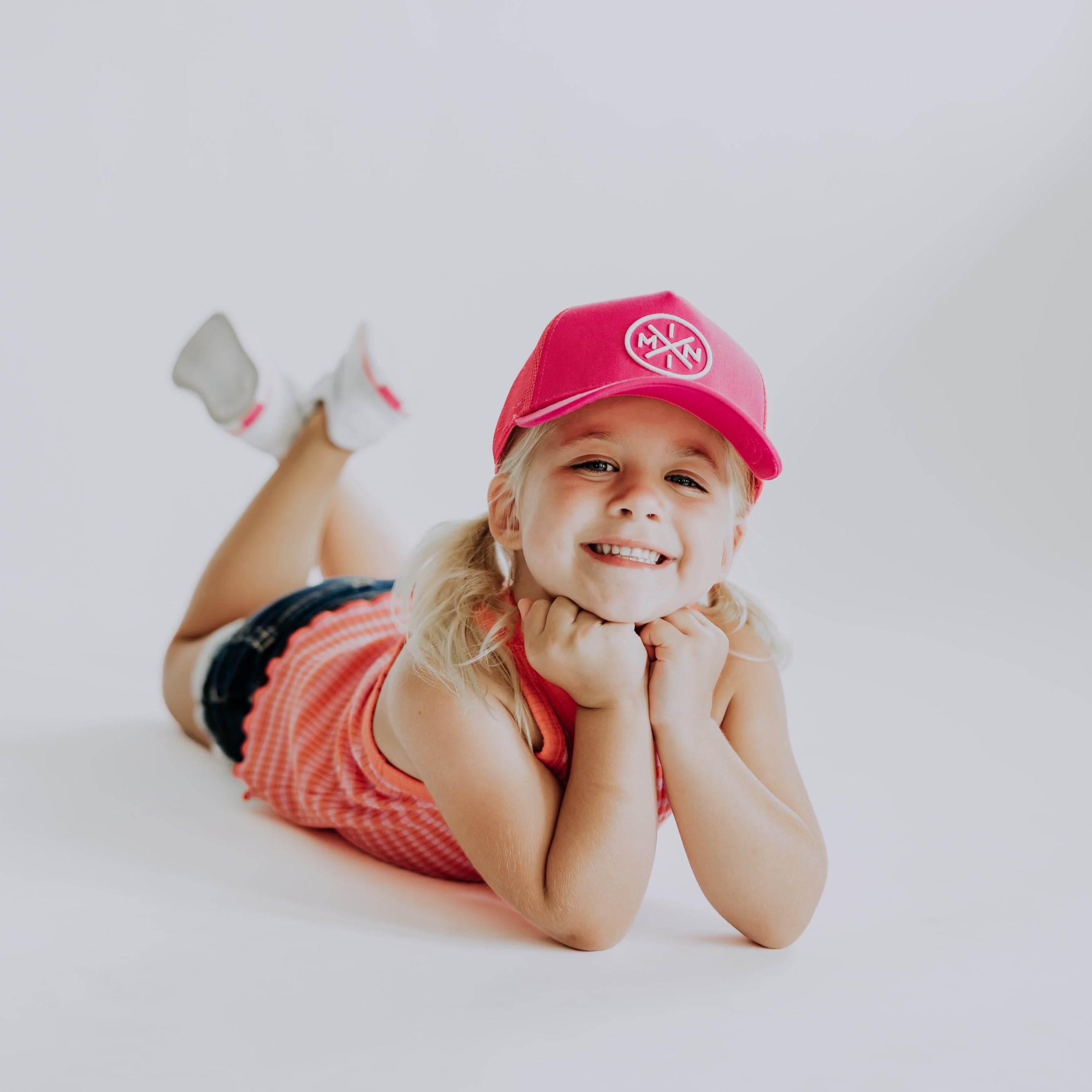 A young child lays on their stomach with legs bent, smiling at the camera. They wear a hot pink Mini X Premium Trucker Hat by Mama X™ Brand, a sleeveless orange top, denim shorts, white socks, and have blonde hair. The background is a plain light color.