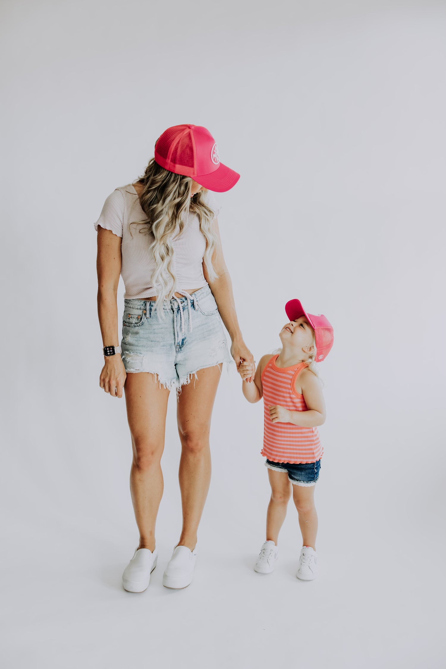 A woman and a young child, both donning Mama X™ Premium Canvas Trucker Hats in hot pink, look at each other and hold hands. The woman is dressed in a pink top with denim shorts, while the child sports a pink-striped tank top paired with denim shorts. Both wear white shoes and stand against a white background.