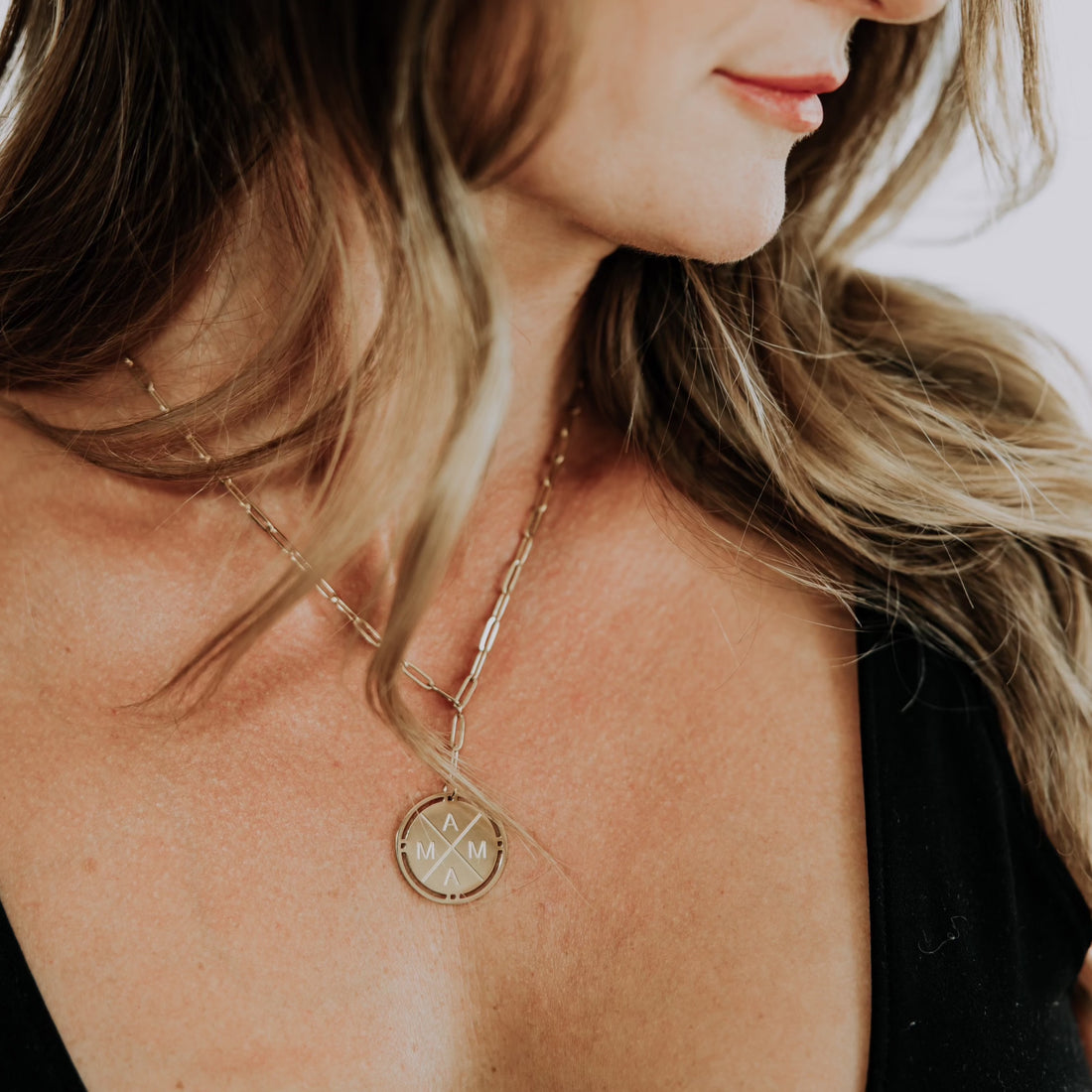 A close-up of a woman with long hair wearing the Mama X™ Gold Necklace by Mama X™ Brand, featuring a round pendant with geometric designs and the letters "M", "A", and "M" visible. The paperclip chain of the necklace gracefully complements her black V-neck top, though her face is partially out of frame.
